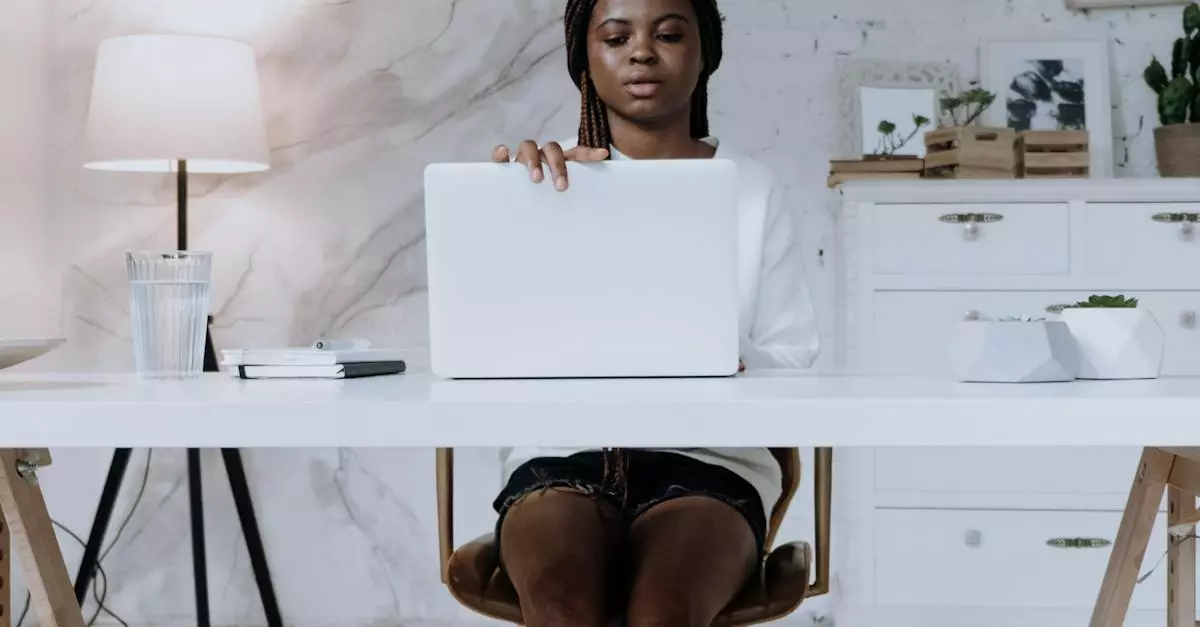 Person Sitting At A Desk Using A Laptop In A Modern Home Office Setting With A Lamp, Plants, And Drinks, Illustrating Digital Marketing And Online Work Trends For 2025.